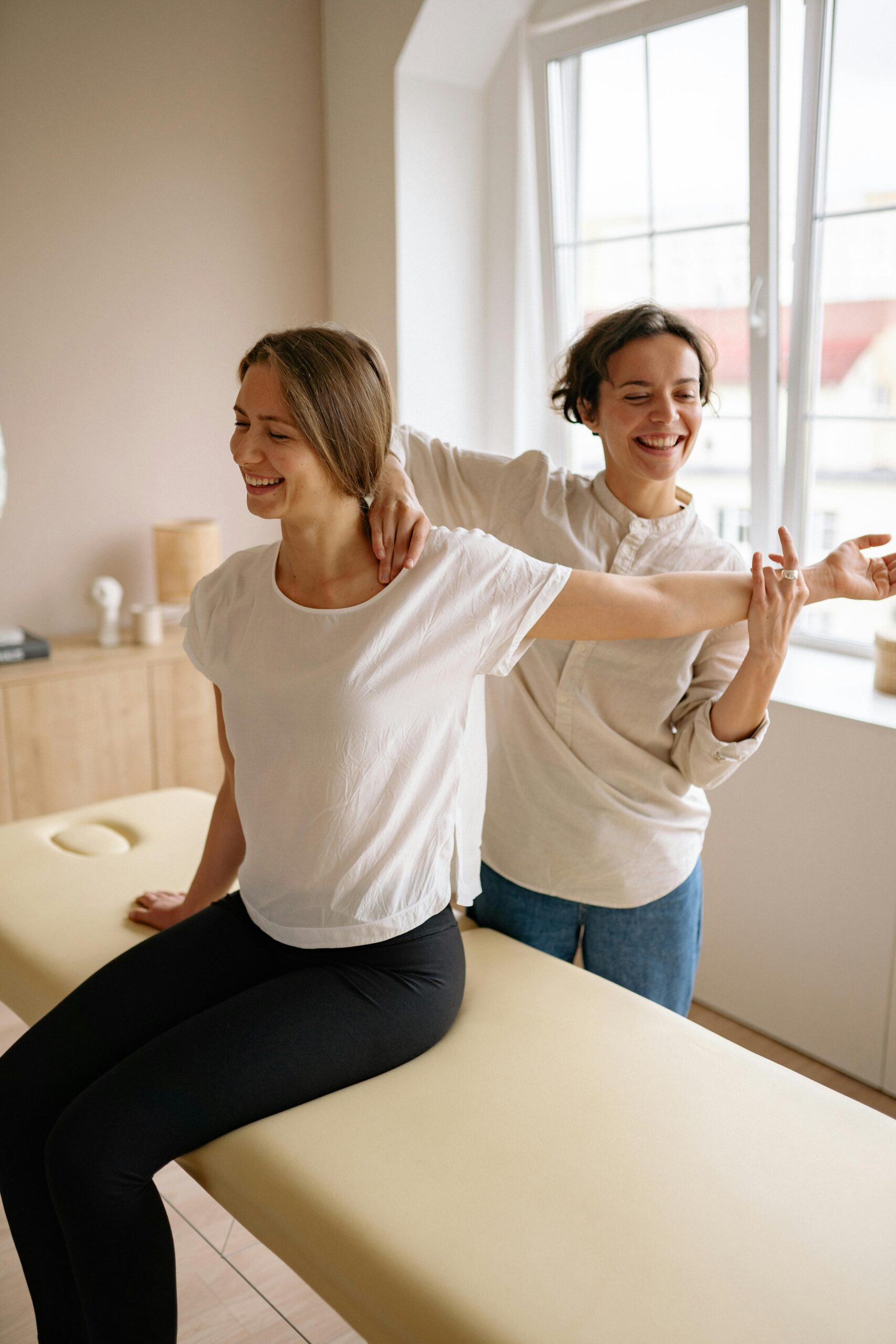 Home A therapist guides a woman in a stretching exercise on a massage table in a clinic setting.